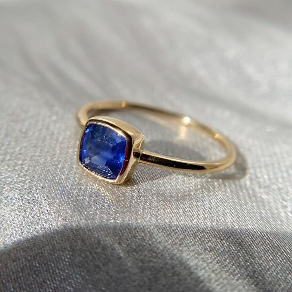 A gold ring with a deep blue rectangular cushion-cut gemstone, possibly a sapphire, set in a bezel setting. The ring is placed on a light-reflective silver fabric background.